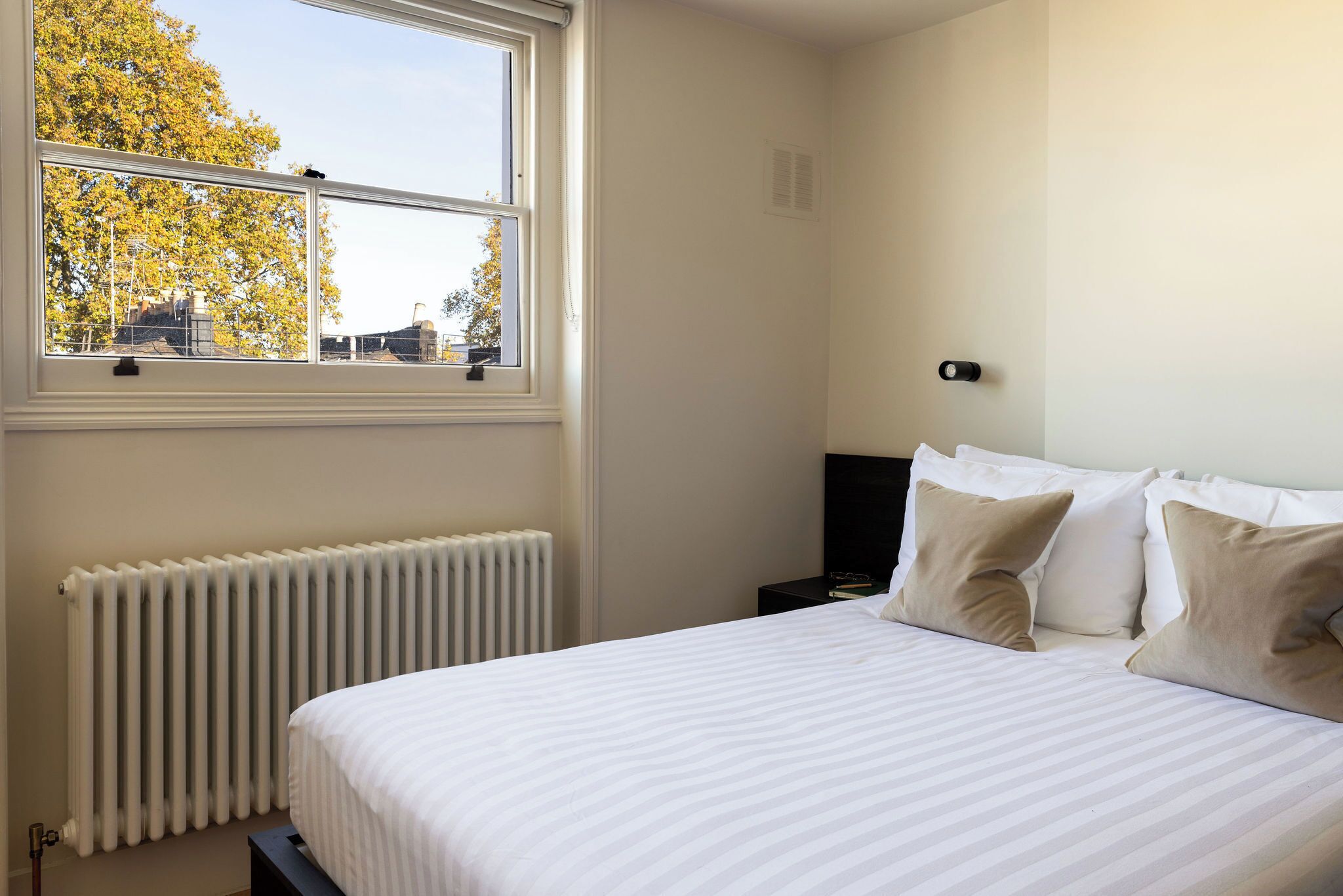 A bedroom with a large bed, a window with a view of trees, a radiator, and a nightstand with a lamp.