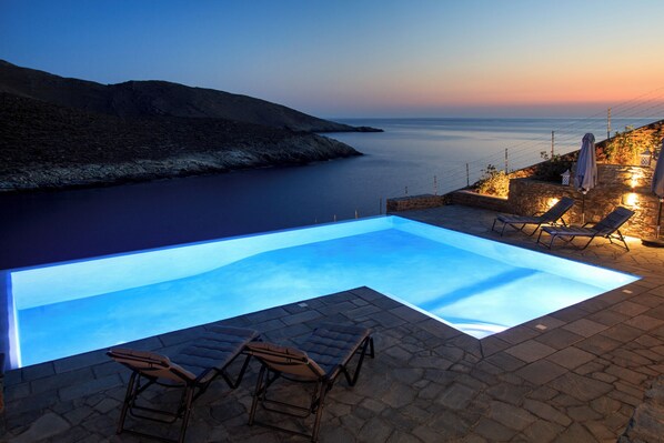 Une piscine avec des chaises longues, un mur en pierre et une vue sur la mer au crépuscule.