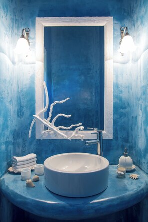 Une salle de bain avec un mur bleu, un lavabo blanc et un miroir. Il y a deux lampes fixées au mur et une branche décorative accrochée au mur.