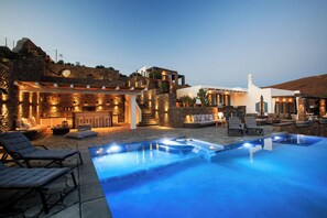 Un espace piscine extérieur luxueux, agrémenté de murs en pierre, de mobilier d’extérieur moderne et d’un bâtiment blanc surmonté d’une tour.