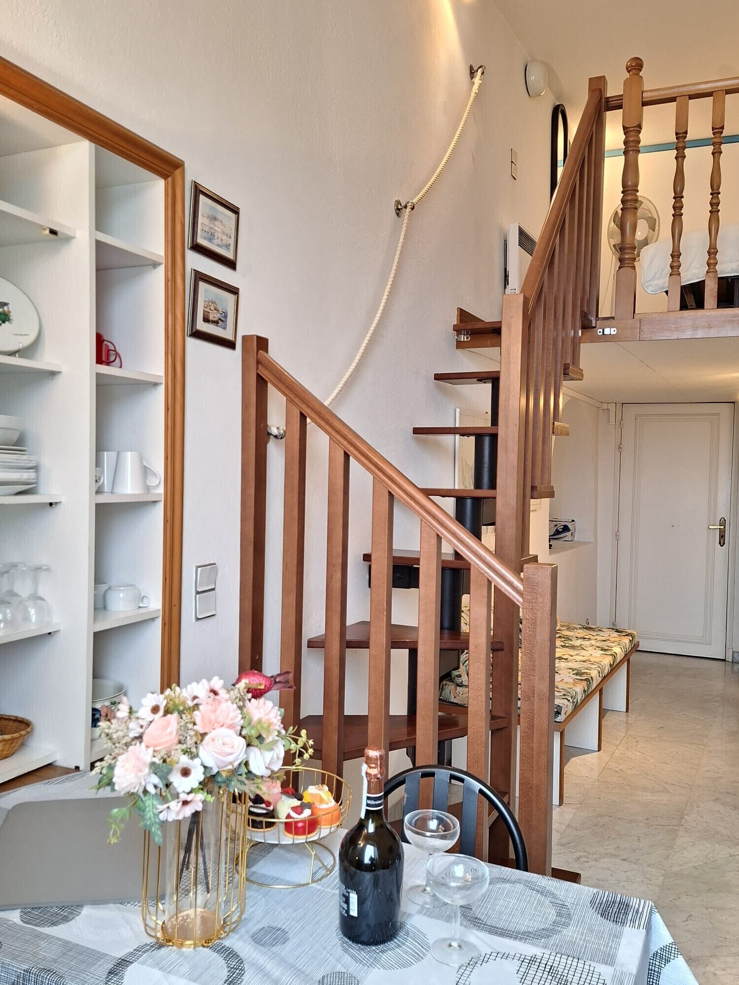 A dining area with a table set for two, a vase of flowers, and a bottle of wine. A staircase with wooden railings leads to an upper level.