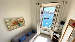 Een woonkamer met een bank, een schilderij en uitzicht op boten op het water door een open raam.
