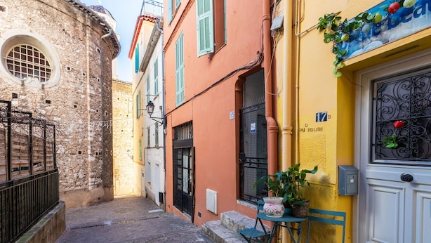 A narrow street with cobblestone pavement, a stone building with a round window, and a yellow building with a door and window shutters. - ESCALINADA II AP4433  by Riviera Holiday Homes (Villefranche-sur-Mer)