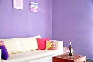 A living room with a white sofa, colorful pillows, a small wooden side table with a wine bottle and glass, and a purple wall. - Marconi Seaside Residence Pool Wifi  (Alba Adriatica)