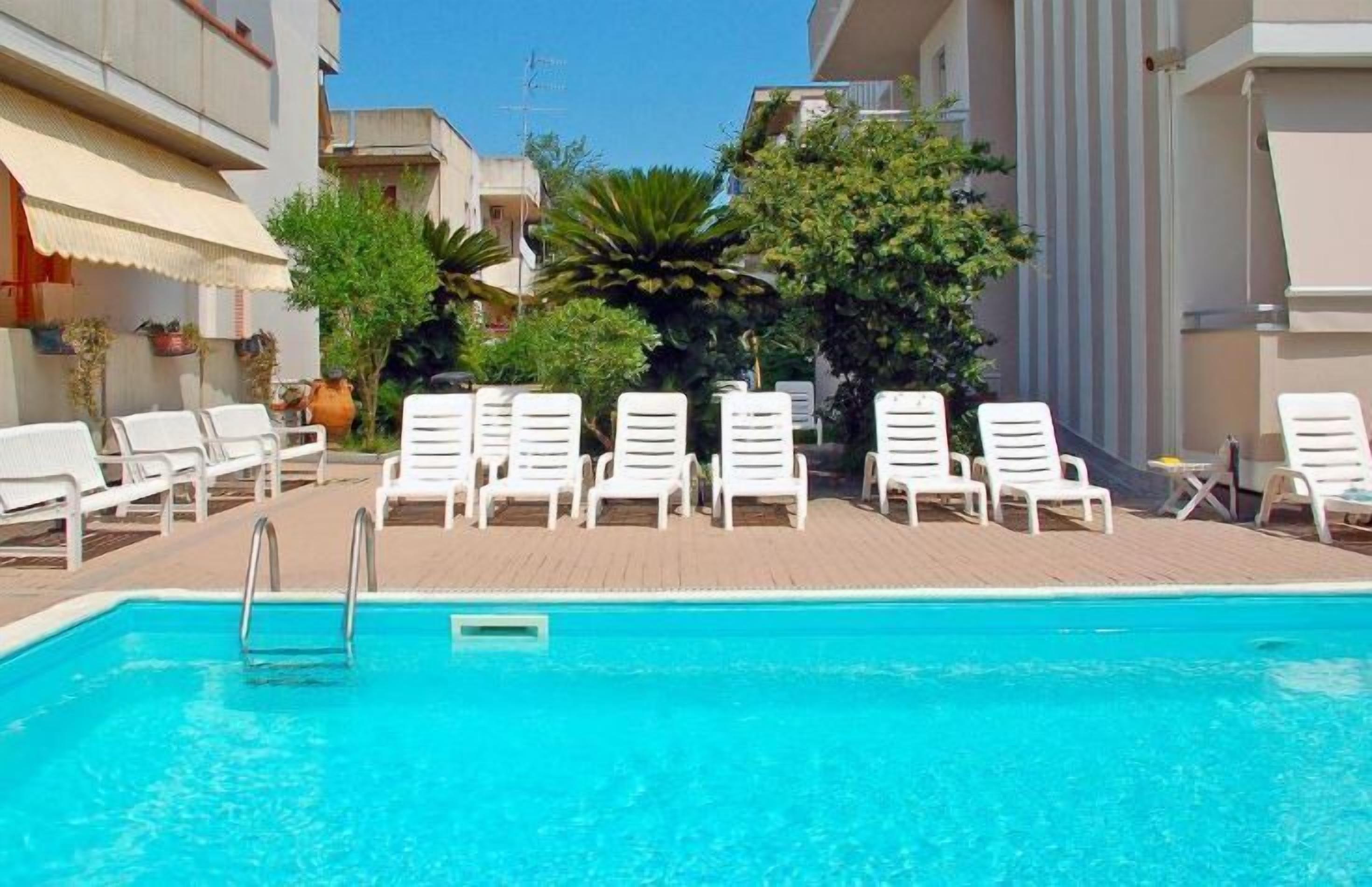 A swimming pool with white lounge chairs arranged around it, surrounded by buildings and greenery.