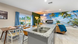 Una cocina moderna con una isla central, un comedor con mesa y sillas, y una sala de estar con un sofá y un mural con motivos de playa.