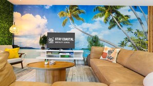 Una sala de estar con un televisor grande, una mesa de centro de madera, un sofá color beige y un mural en la pared que representa una playa.