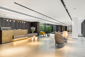 A modern reception area with a wooden reception desk, seating with cushions, and a view of greenery outside. - MAMAD Luxury 3BR Next to Geula Beach by FeelHome (Tel Aviv - Jaffa)
