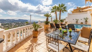 Une terrasse avec une table en verre, des chaises, des plantes en pot et une vue sur la mer et les montagnes.