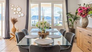 Un coin repas avec une table en verre, des chaises à revêtement gris, un vase de fleurs et une vue sur l’extérieur grâce à de grandes fenêtres.