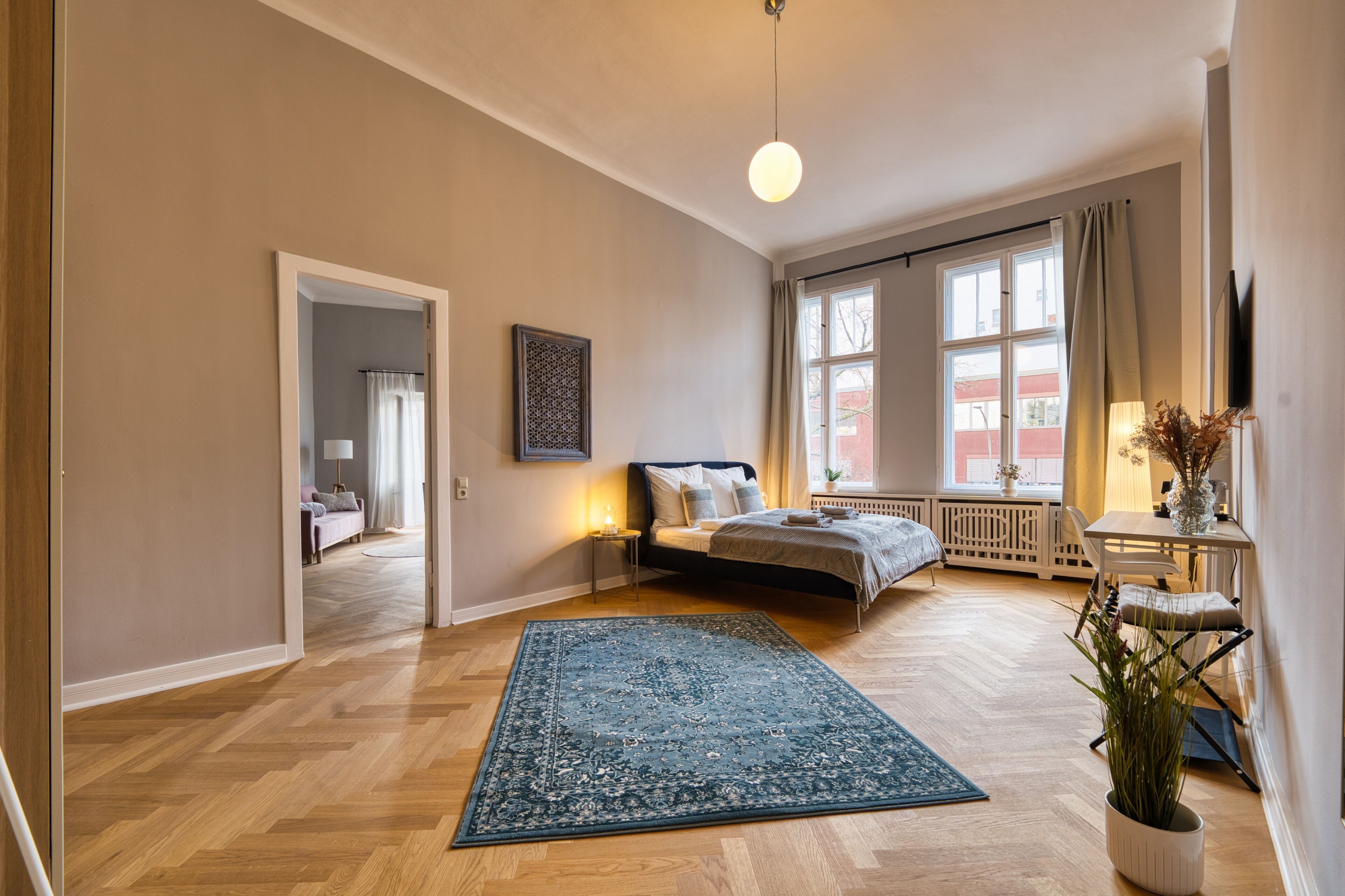A spacious bedroom with a large bed, a bedside table with a lamp, a window with curtains, and a view of the outside.