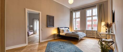 A spacious bedroom with a large bed, a bedside table with a lamp, a window with curtains, and a view of the outside.