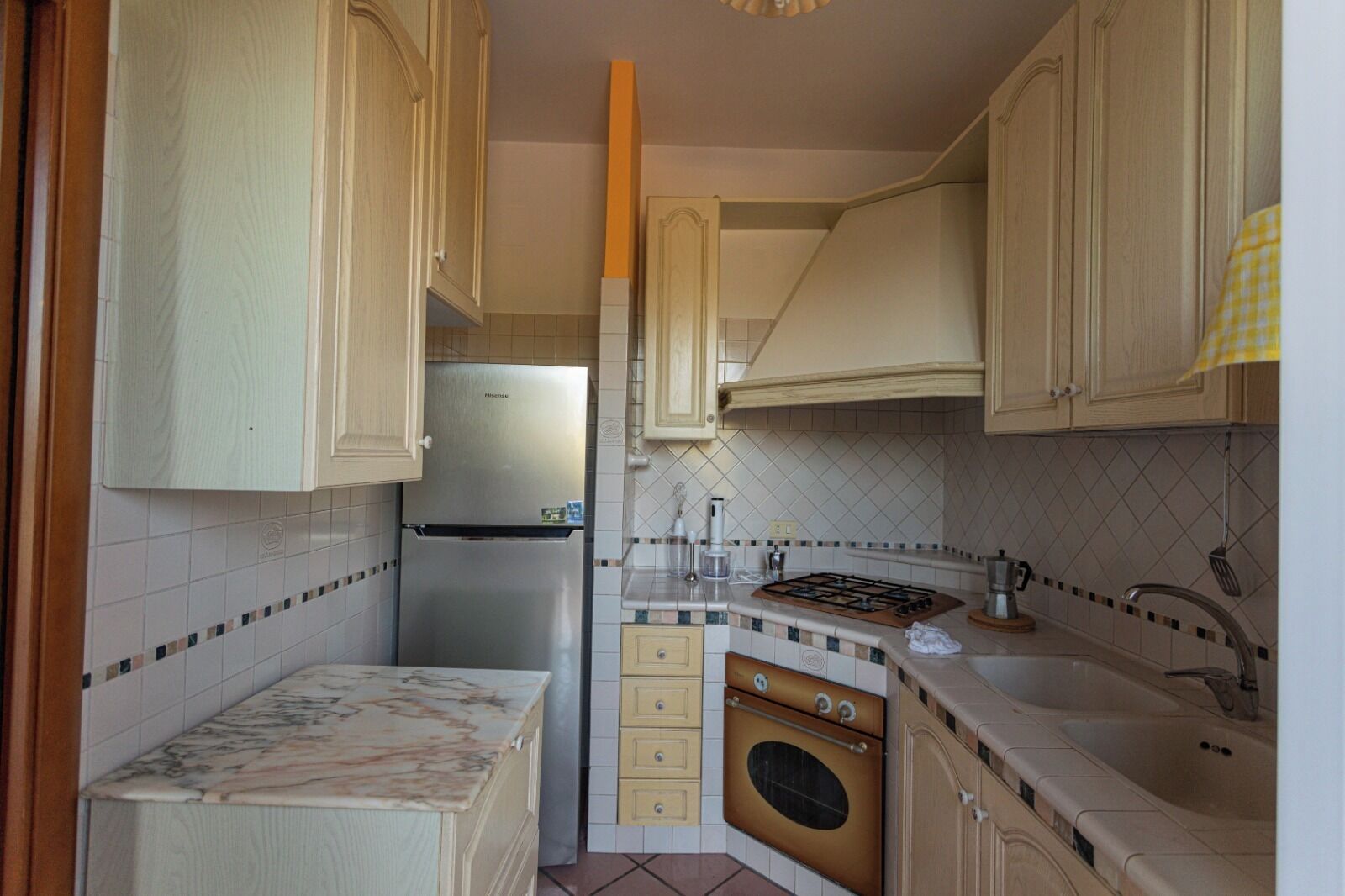 A compact kitchen with light wooden cabinets, a stainless steel refrigerator, a gas stove, and a built-in oven.