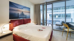 A hotel room with a large bed, a desk, a chair, a window with a city view, and a painting on the wall. - Waterfront Seaside in Genova (Genoa)