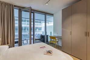 Camera d'albergo con un letto, una sedia, una scrivania e un armadio. Si vede una finestra con tende e vista su un edificio.