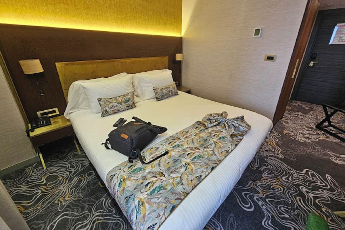 Quarto de hotel com uma cama grande, duas almofadas, um cobertor, uma mala e uma mesa de cabeceira com luminária.