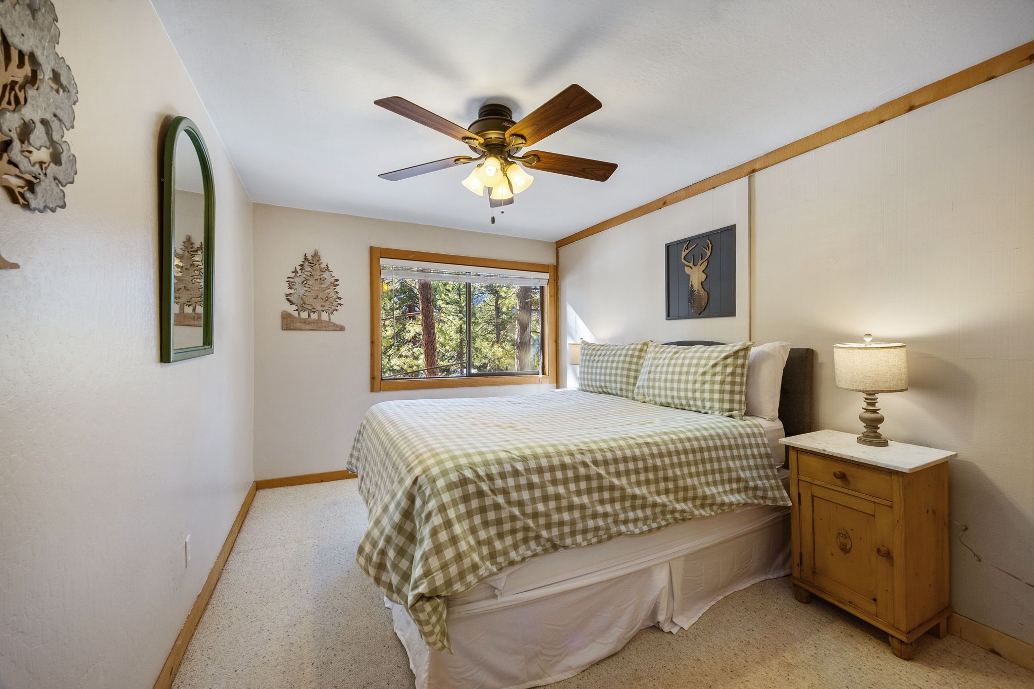A bedroom with a bed, a nightstand, a ceiling fan, a window with a view of trees, and wall art.
