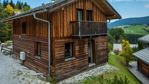 Una cabaña de madera con balcón, rodeada por un camino de grava y una zona ajardinada.