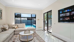 A living room with a sectional sofa, a round coffee table, a large TV screen, and a view of a swimming pool. - 6BR Lower Mitcham home, pool, cinema, near CBD (Lower Mitcham)