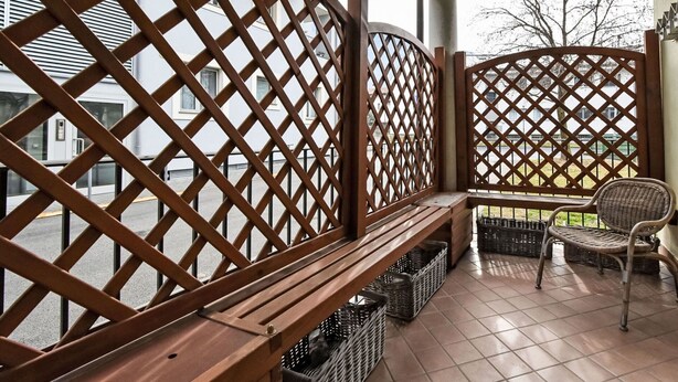 Ein Balkon mit Holzgitter, einer Bank, Korbstühlen und Körben am Boden.