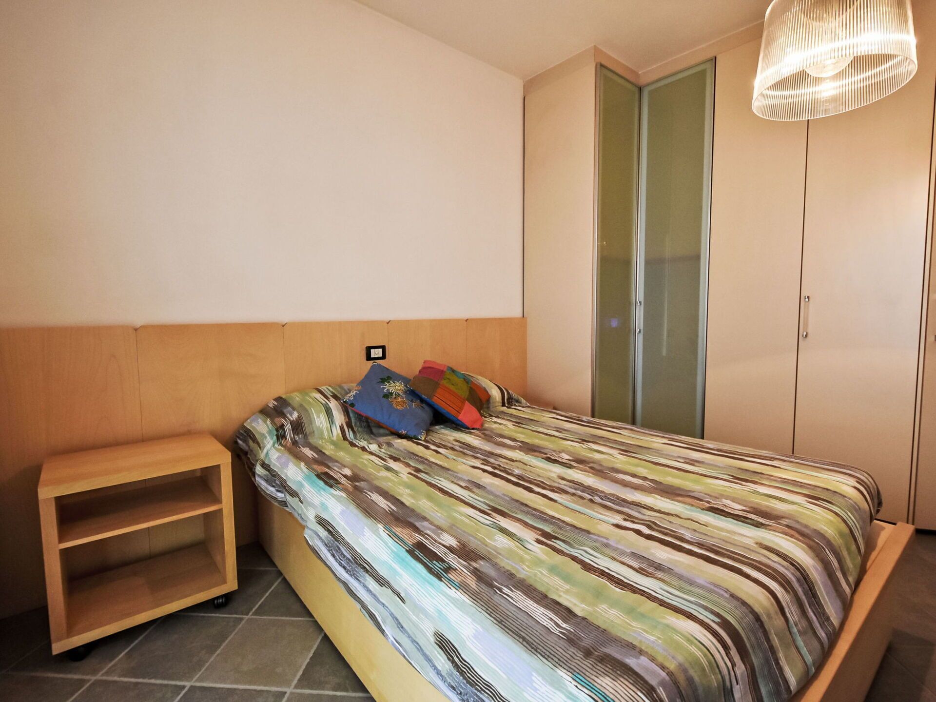Un dormitorio con una cama, una mesita de noche de madera, un armario y una lámpara colgante.