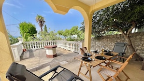 A covered outdoor dining area with a table set for four, surrounded by chairs, a stone wall, and a view of greenery and a palm tree. - Kuća za odmor Fay (Crikvenica)