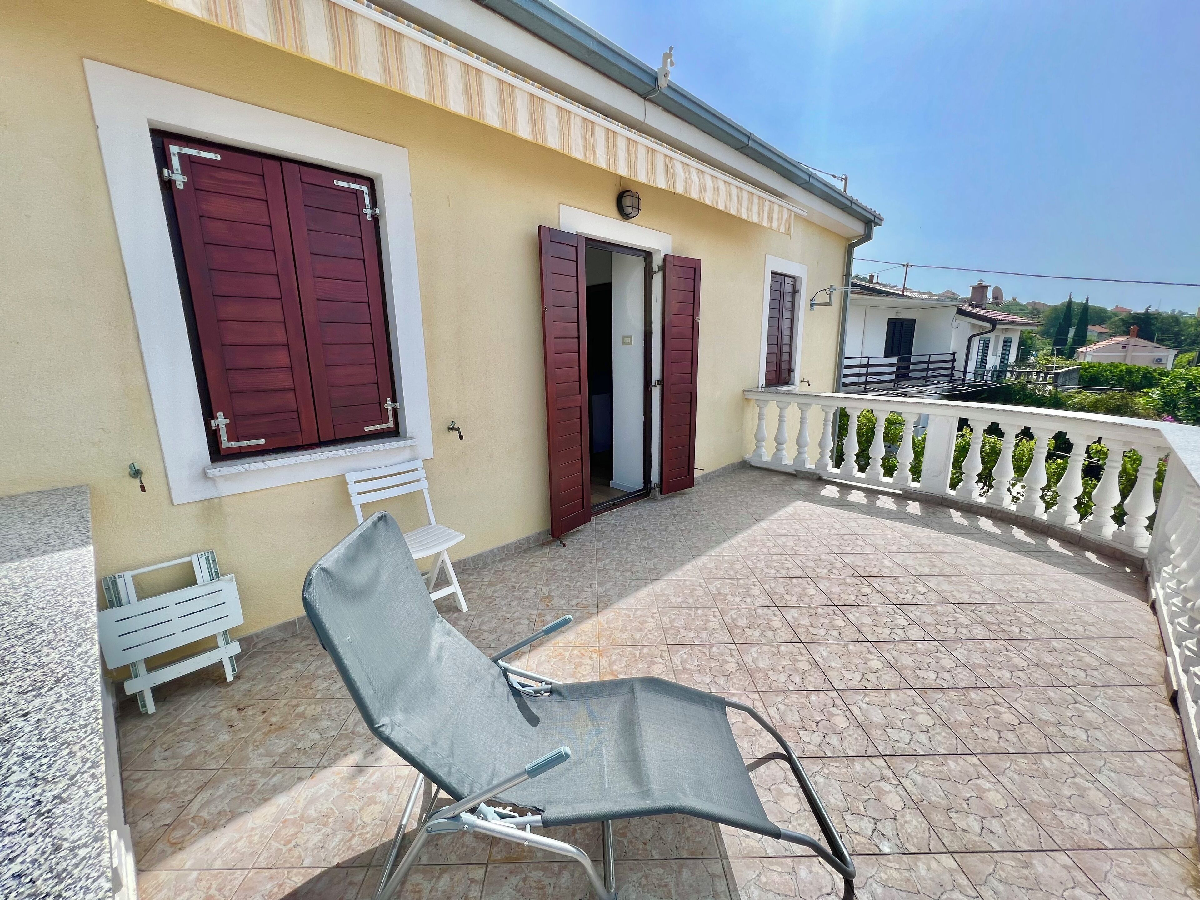 Un balcon avec une balustrade blanche, deux chaises grises et une porte avec des volets rouges.