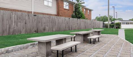 An outdoor patio area with concrete tables and benches, a wooden fence, and a grassy lawn.