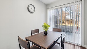 A dining area with a wooden table, four chairs, a clock on the wall, and a view through large windows.
