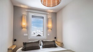 A bedroom with a large bed, two wall-mounted lamps, a window with a view of a snowy mountain, and a woven ceiling light fixture. - Cozy apartment at the slopes with balcony (Val Thorens)