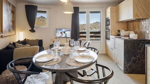 A dining area with a round table set for four, a kitchen with wooden cabinets, and a view of mountains through the windows. - Cozy apartment at the slopes with balcony (Val Thorens)