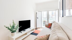 A modern living room with a flat-screen TV on a white stand, a potted plant, a beige sofa, and a large window offering a view of a balcony. - Seaside Modern Flat II at Cascais (Cascais)