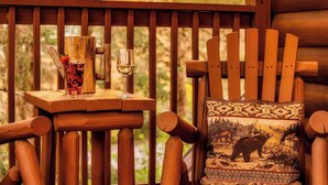A wooden deck with a table holding a glass of white wine and a drink with a straw. A wooden chair with a patterned cushion is also visible.