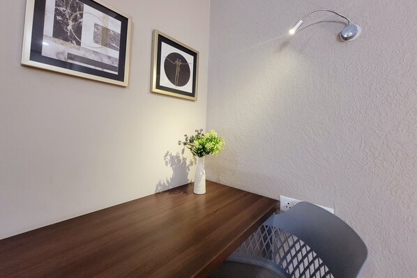 Un escritorio de madera con un jarrón blanco lleno de flores, dos cuadros en la pared y una lámpara fijada a la pared.
