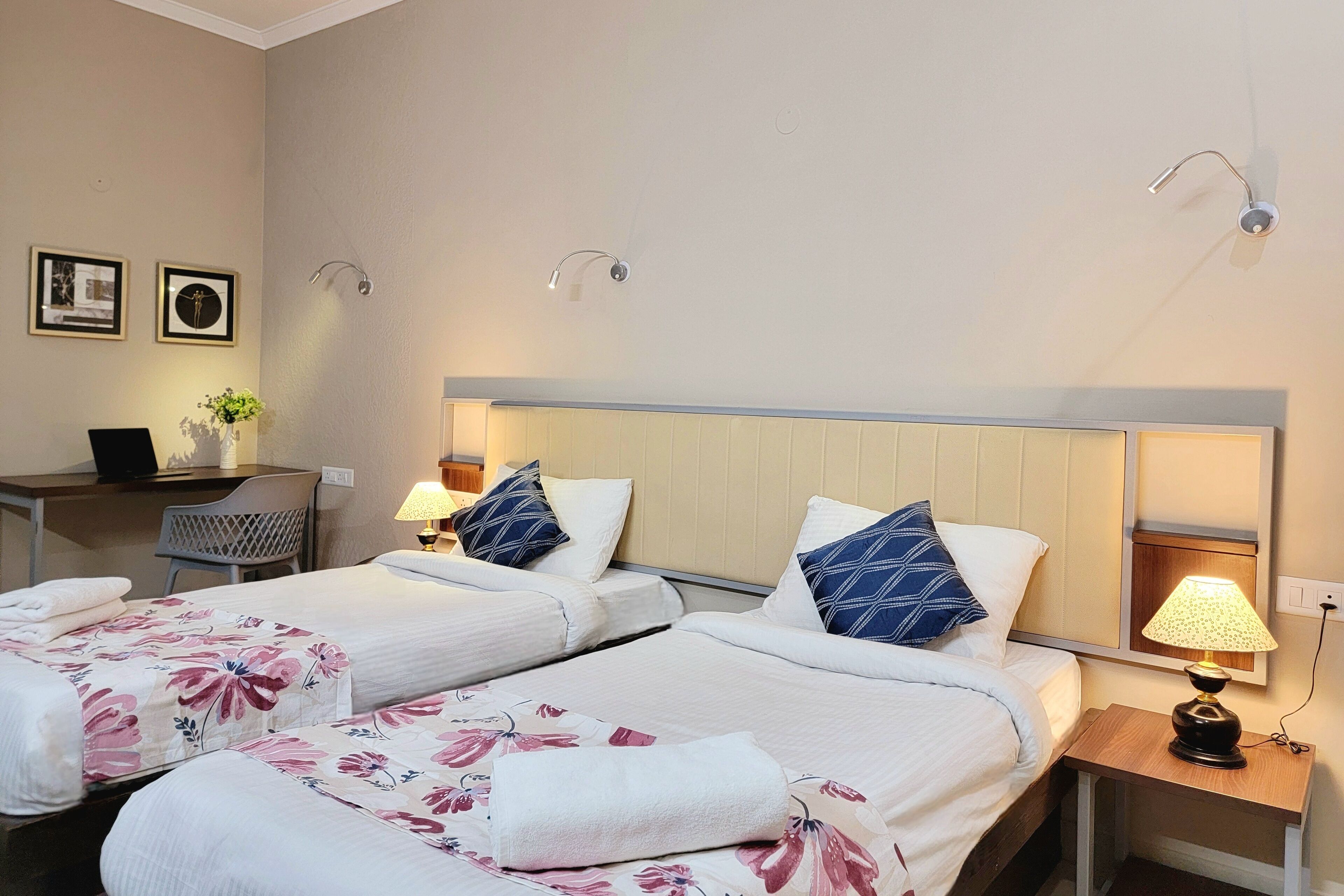 A hotel room with two beds, a desk with a laptop, a small table with a lamp, and wall-mounted lights.