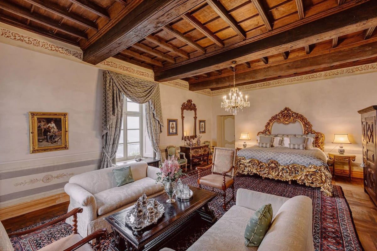 Ein traditionell eingerichteter Raum mit Holzbalkendecke, einem großen Bett mit aufwändiger Bettwäsche, einer Sofagarnitur und einem Chandelier.
