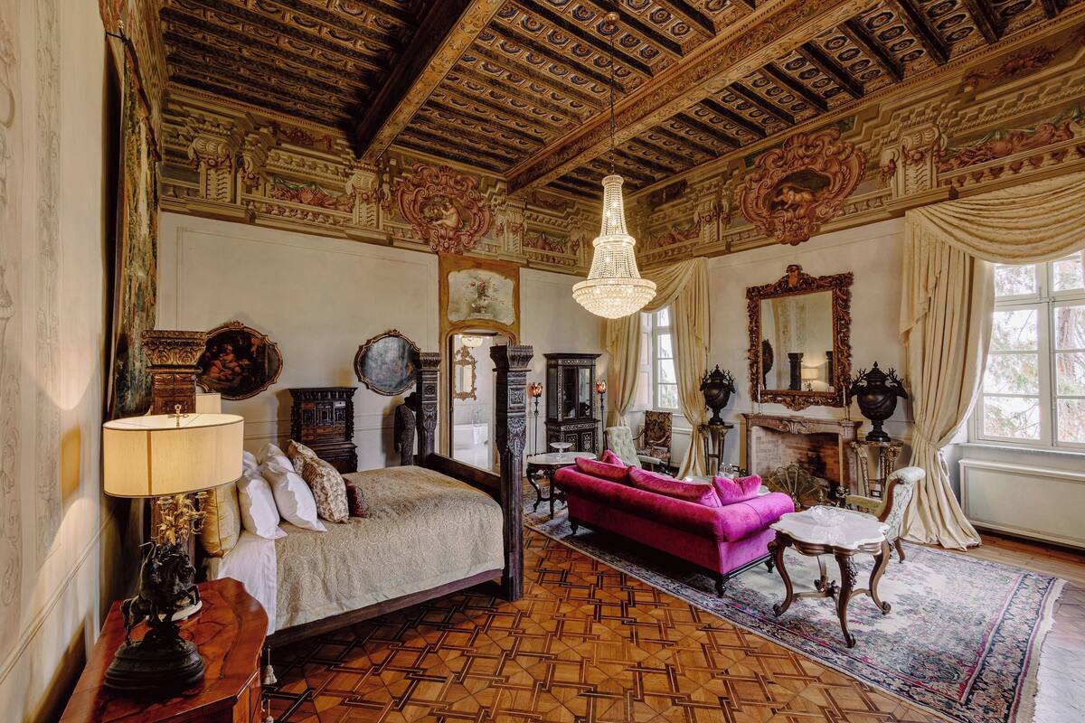 Ein Zimmer mit einem Bett, einem rosafarbenen Sofa, einer Chandelüster und aufwendigen Deckenverzierungen.