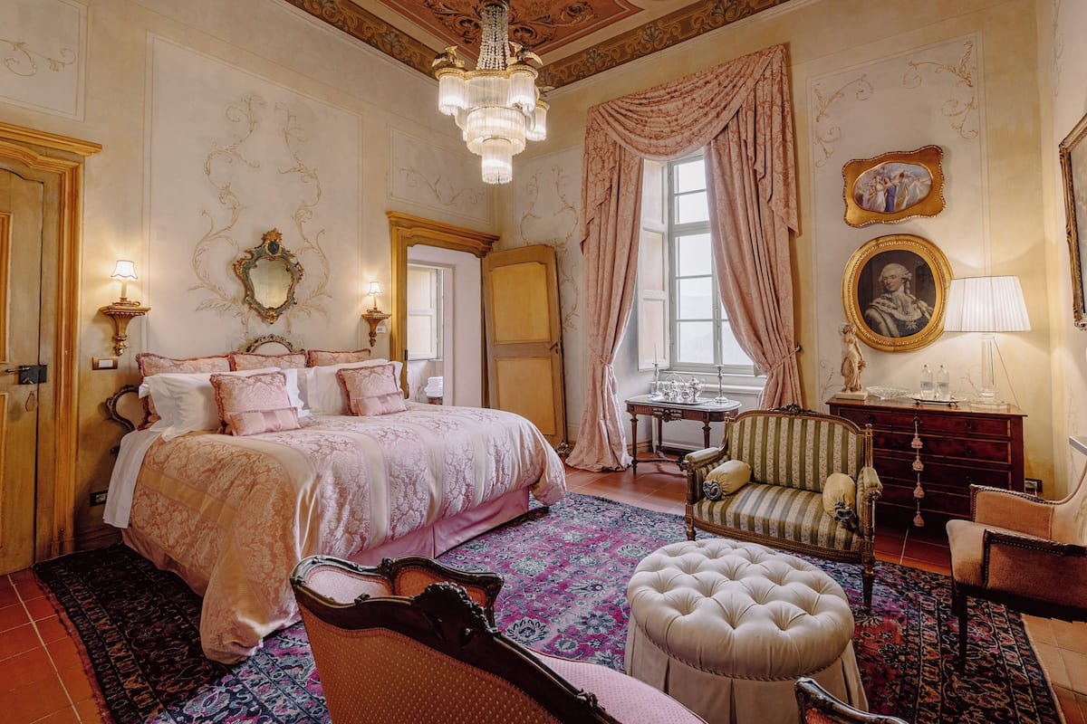 Ein klassisches Schlafzimmer mit einem großen Bett, einer Sitzecke mit Sessel und Hocker, einem Schminktisch und einer Chandelüster.