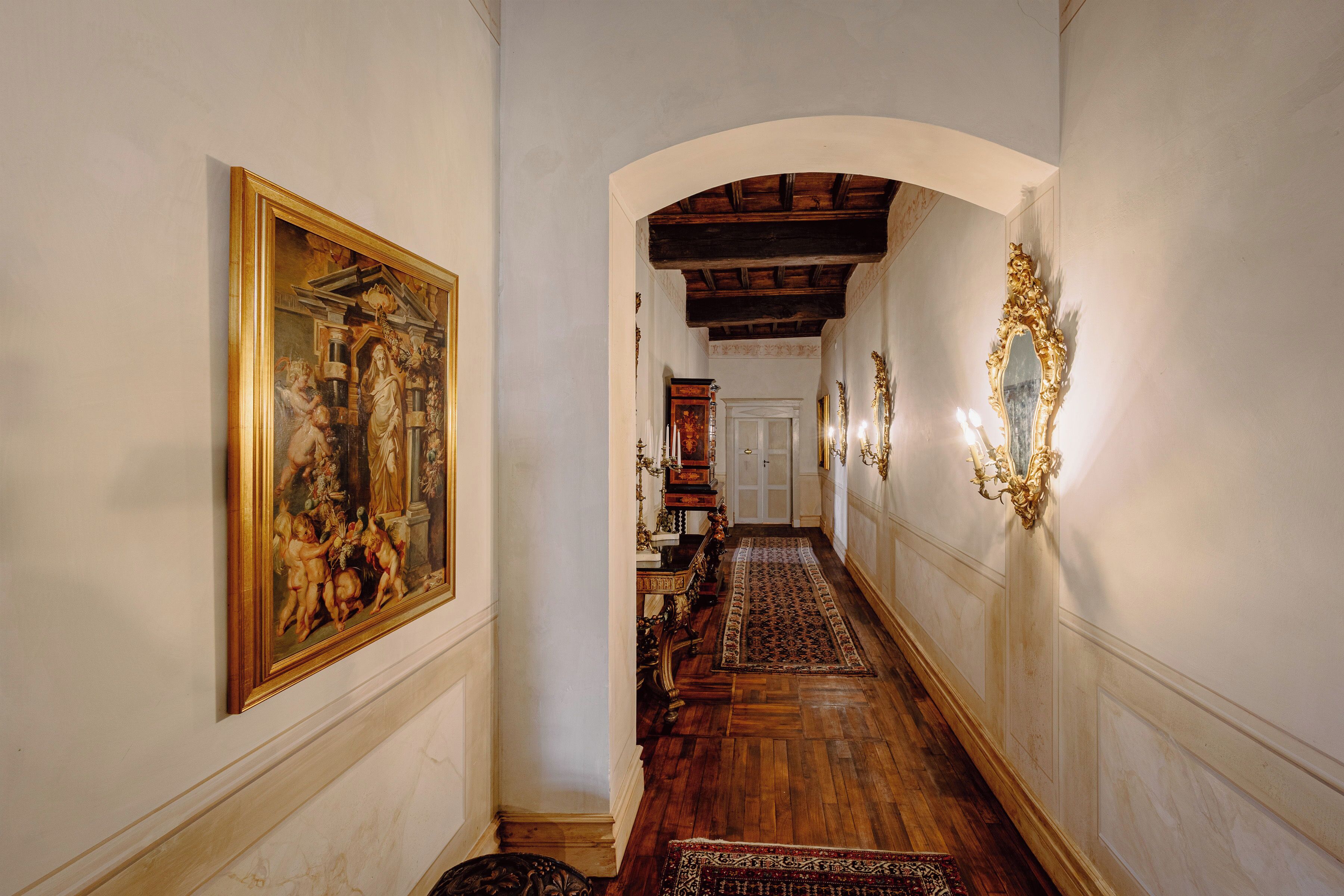 A hallway with a wooden floor, a large framed painting on the left, and a decorative wall sconce on the right.