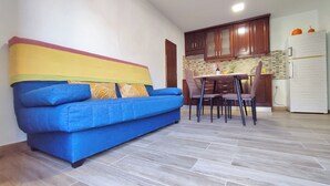 A living room with a blue sofa, a dining table with chairs, wooden cabinets, and a white refrigerator.