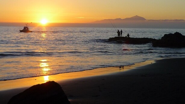 A beach at sunset with a boat, people fishing, and a mountain in the background.