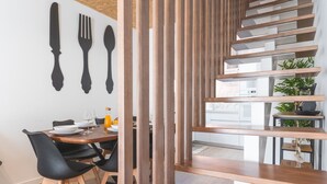 Uma zona de refeições moderna com uma mesa de madeira, cadeiras pretas e uma cozinha visível através de uma divisória de madeira.