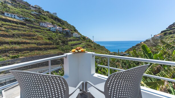Uma varanda com duas cadeiras, uma mesa com frutas e vista para o mar e casas na encosta.