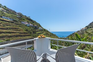 A balcony with two chairs, a table with fruit, and a view of the sea and hillside houses.