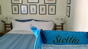 Quarto de hotel com uma cama, um cobertor azul e um travesseiro com a palavra "Sicilia" bordada. Há quadros na parede acima da cama.