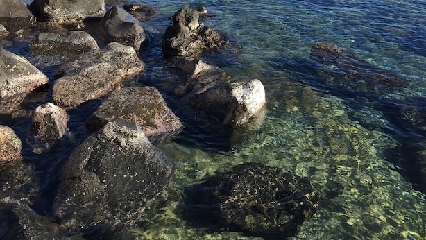 Uma costa rochosa com águas cristalinas, uma praia de areia e barcos ao fundo.
