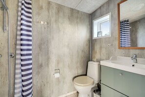 A bathroom with a concrete wall, a white toilet, a sink with a mirror, and a striped shower curtain. - Bjålumvej 17 (Norre Nebel)