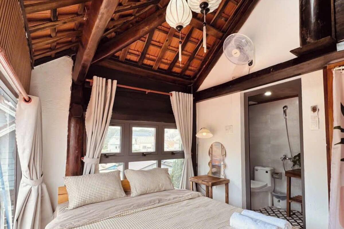 Camera da letto con soffitto in legno, un letto con copriletto a quadretti, un comodino, una lampada e una finestra con tende.