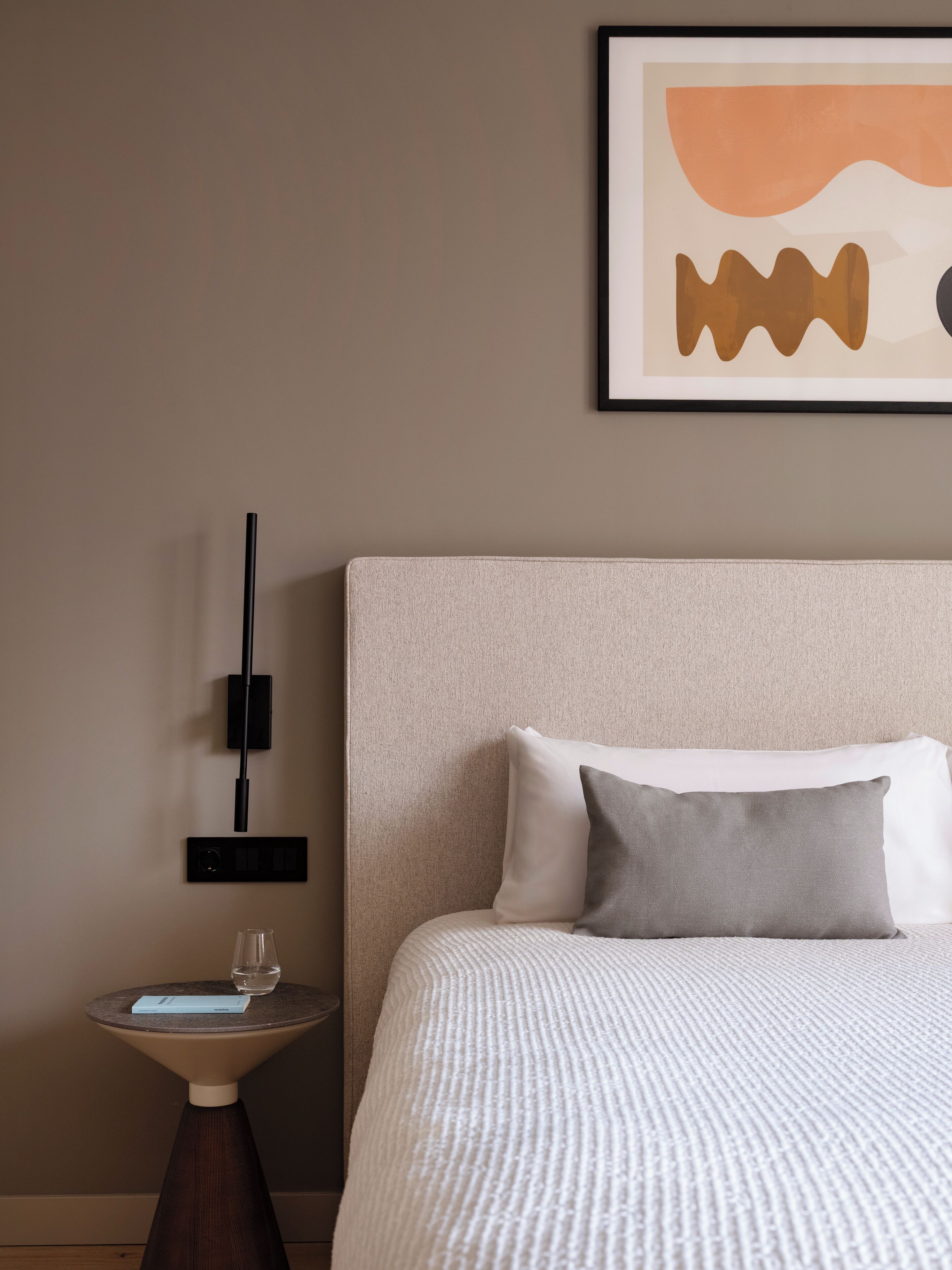 A neatly made bed with a textured white duvet, a grey pillow, and a white pillow. A small bedside table with a glass of water and a blue card is visible. A framed abstract artwork hangs on the wall above the bed.
