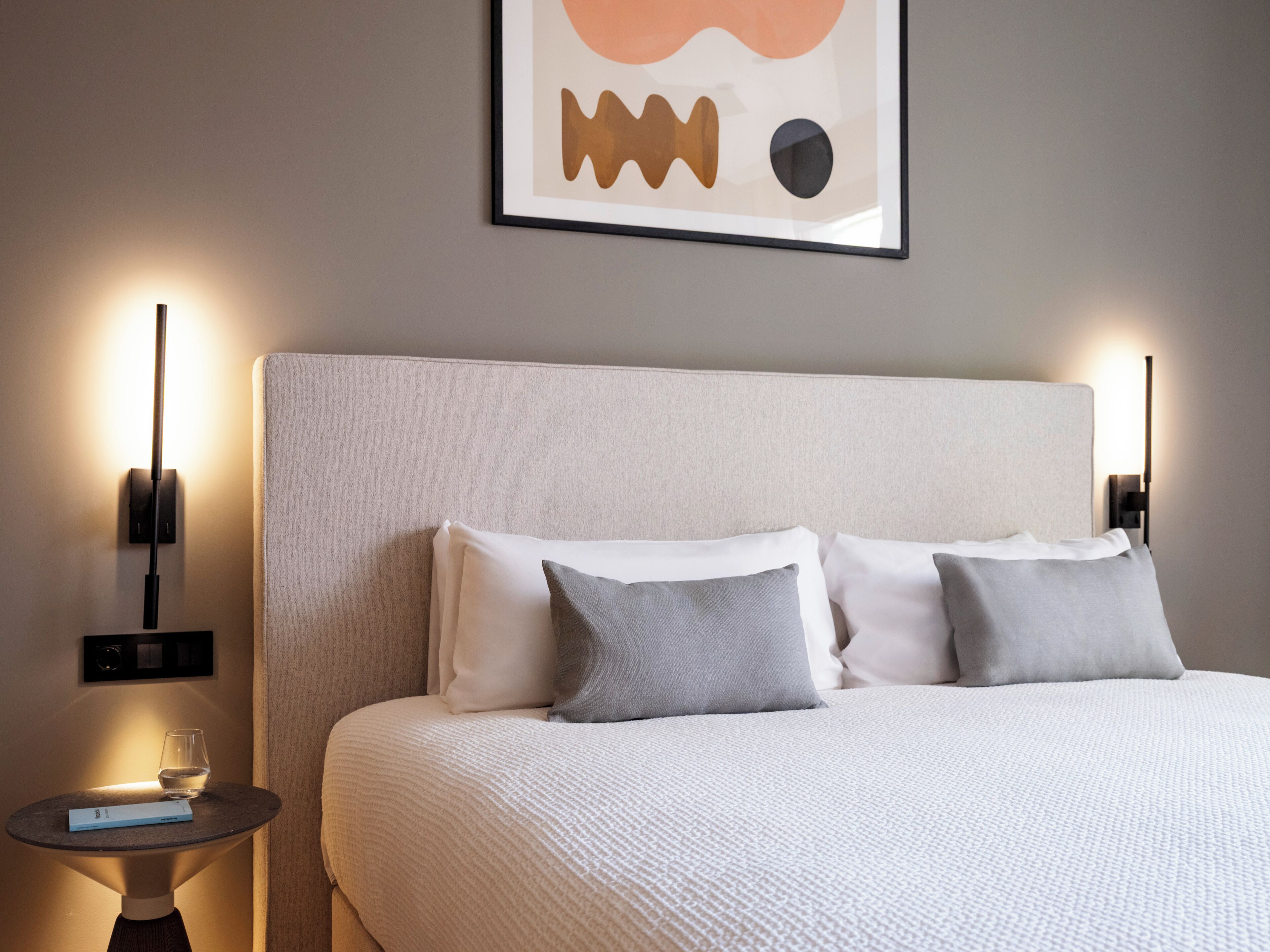 A neatly made bed with white and grey bedding, a bedside table with a glass of water and a book, and two wall-mounted lamps.